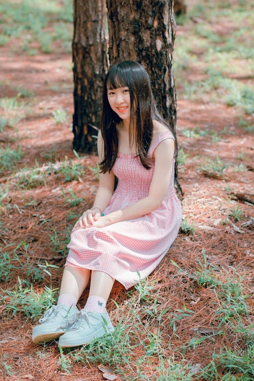 Girl's Pink Sleeveless Dress Sits Beside Black Tree Trunk at Daytime