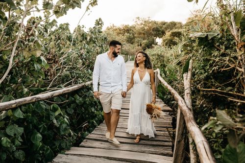 Free Happy newlywed couple holding hands and walking on a scenic wooden bridge surrounded by lush greenery. Stock Photo