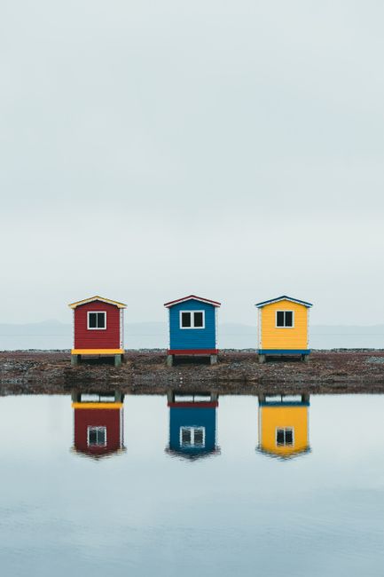 Three vibrant fishing sheds in red, blue, and yellow reflecting on a calm water surface.