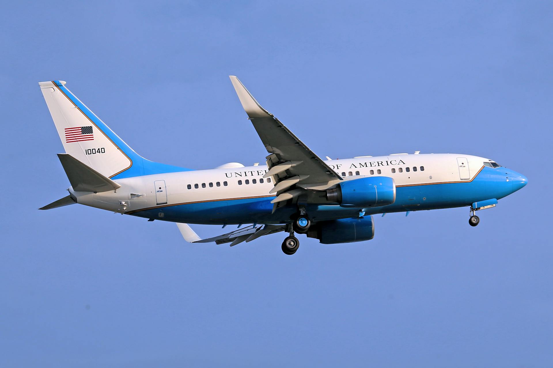 A US government airplane soaring through a clear blue sky, displaying the American flag prominently.