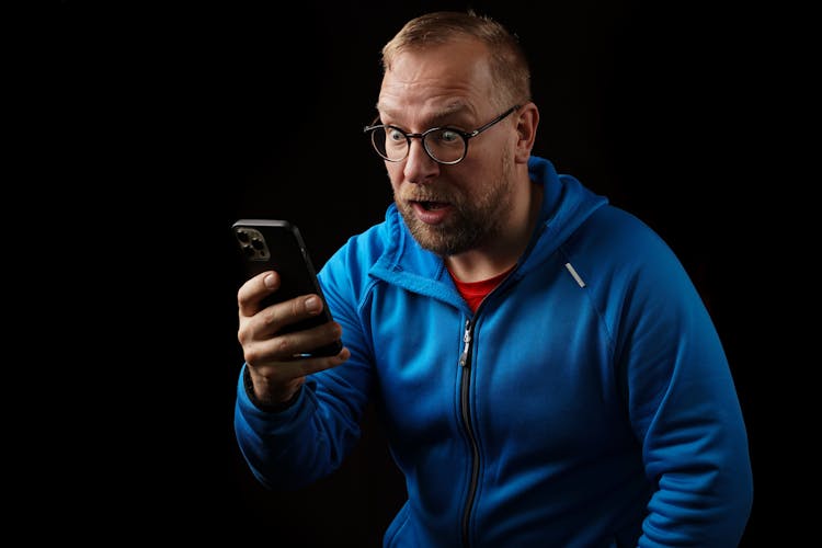 A Man In Blue Zip Up Jacket Looking Shocked While Looking At The Screen Of A Cellphone