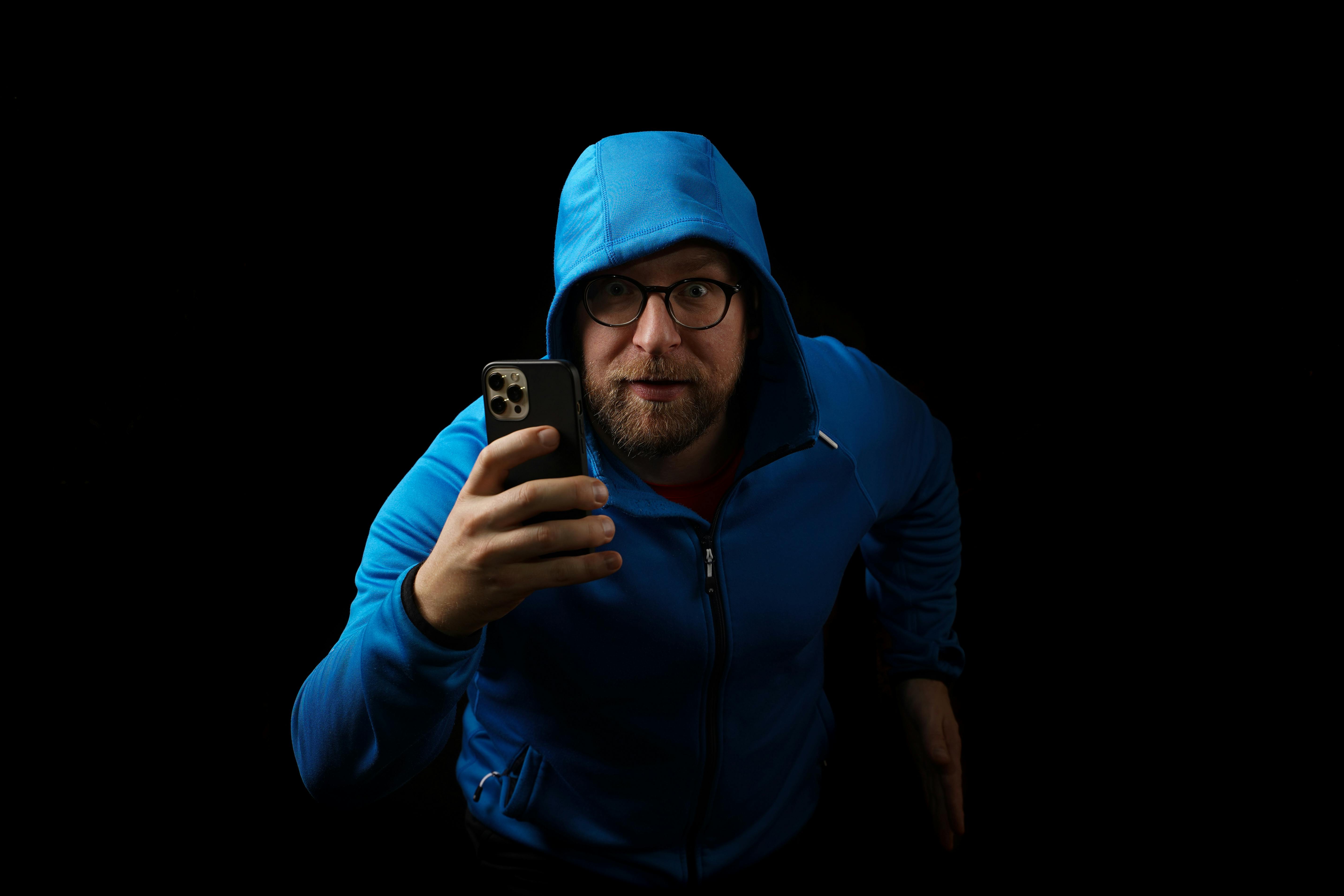 A man in a blue hoodie, wearing eyeglasses, holds a smartphone against a black background in a studio setting.
