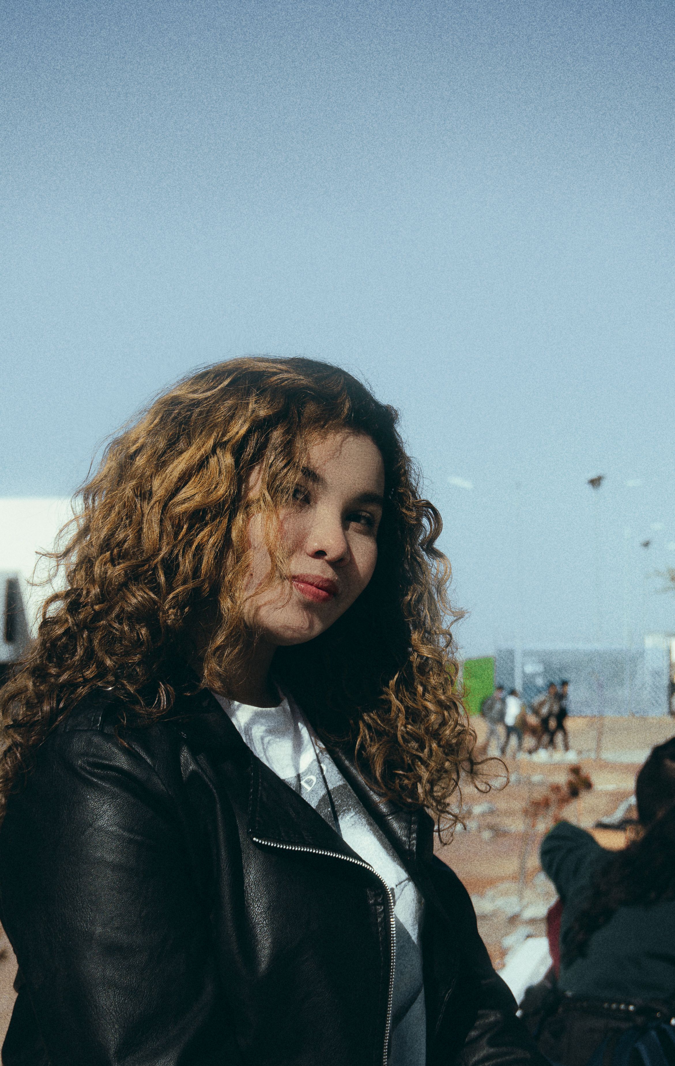 Stylish young woman with curly hair in a black leather jacket, posing outdoors with a confident smile.
