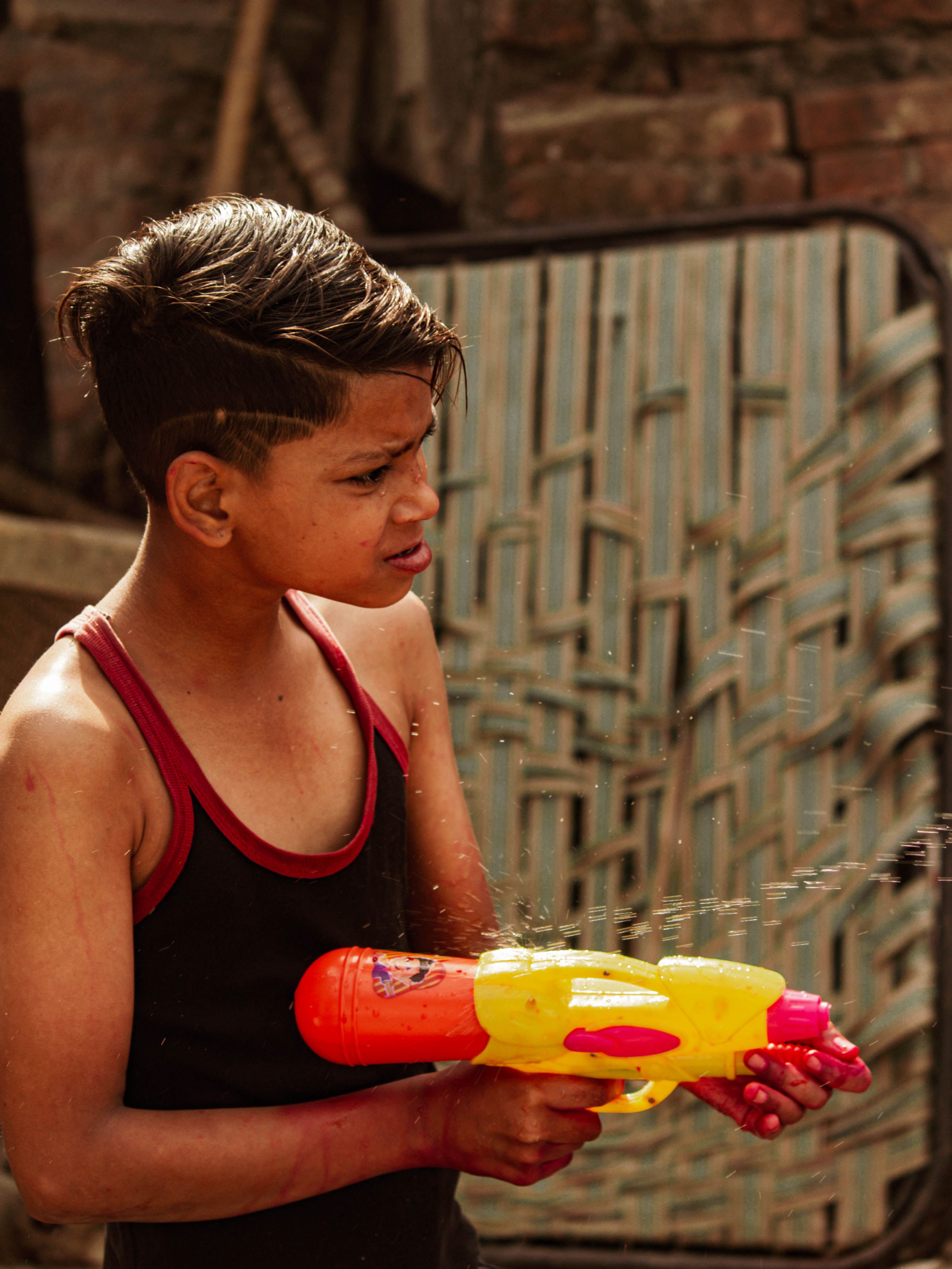 A young boy in a black tank top playing joyfully with a colorful water gun outdoors.