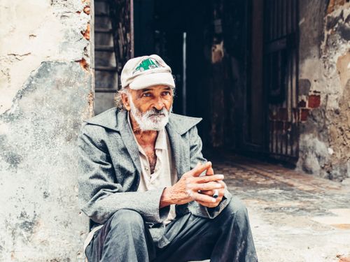 Free An elderly man with a beard and cap sits outdoors in an urban area, exuding a contemplative mood. Stock Photo