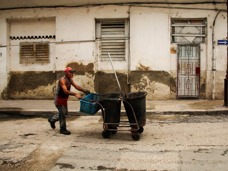 An Elderly Man Pushing A Cart With Trash Cans And A Plastic Crate