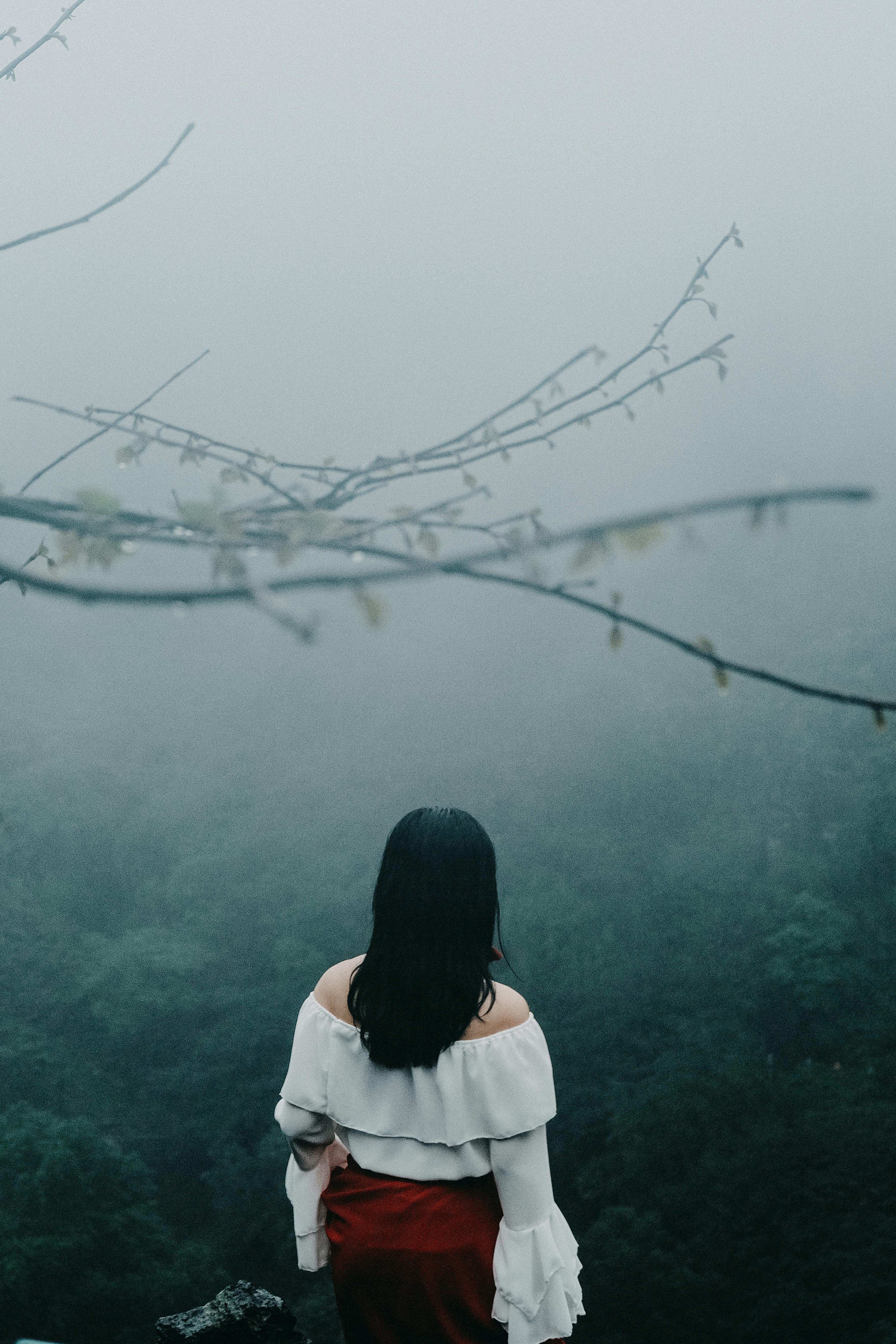 A woman in an off-shoulder dress stands facing a misty landscape, conveying solitude.