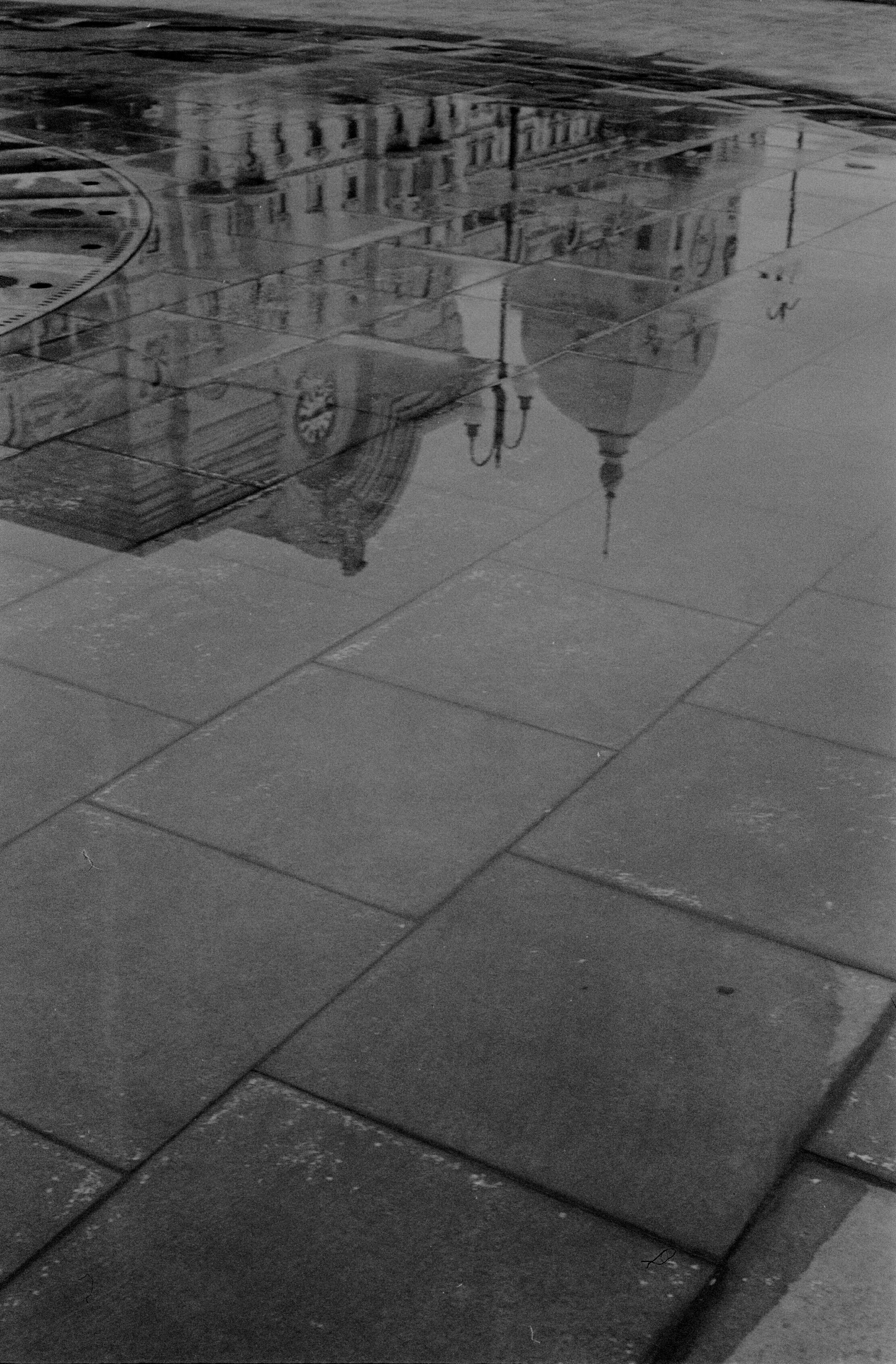Black and white photograph capturing a historic building's reflection in a rain puddle on a city street.