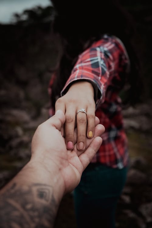 A Person in Plaid Long Sleeves Wearing Engagement Ring while Holding a Tattooed Person Hand