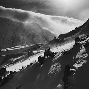 Dramatic black and white photo of skiers descending a sunlit snowy mountain slope with clouds.
