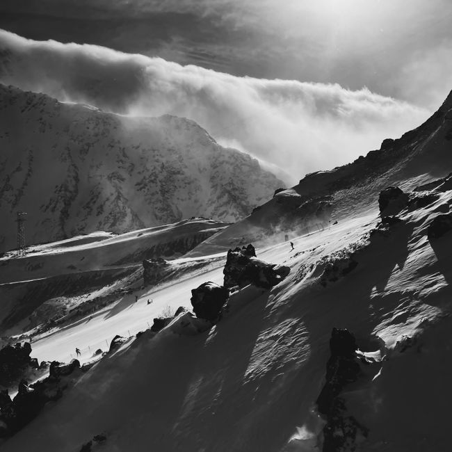 Dramatic black and white photo of skiers descending a sunlit snowy mountain slope with clouds.