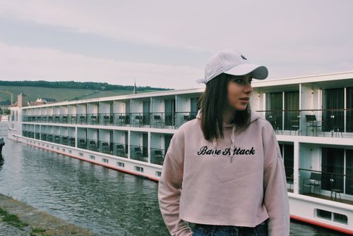 Free A young woman in a cap and hoodie stands by a canal in Würzburg, Germany, with a riverboat in the background. Stock Photo