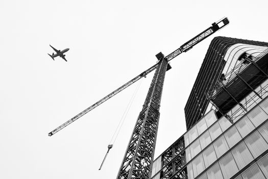 A striking grayscale image of a modern building under construction with a tower crane and airplane in the sky.
