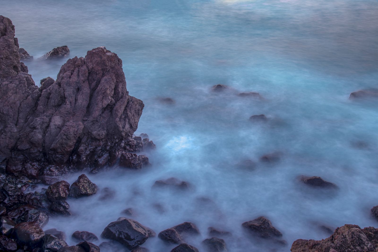 Dark coastal rocks surrounded by soft blue water in a long exposure.