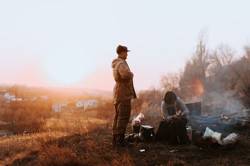 Free Two people enjoy a campfire on a hill at sunset, creating a warm, outdoor experience. Stock Photo