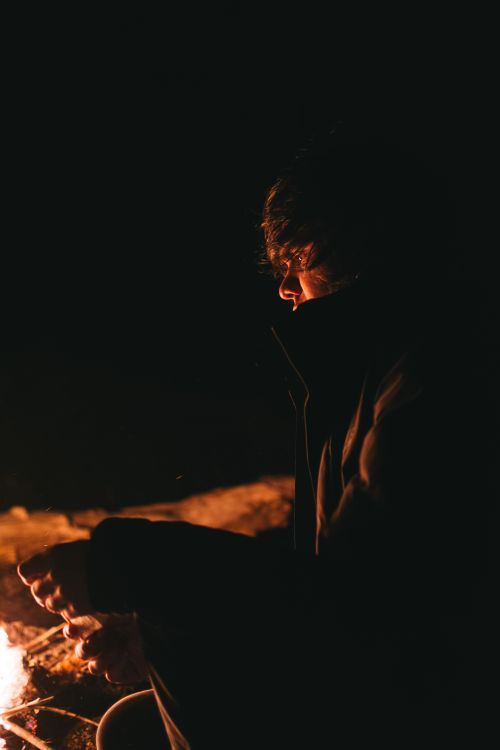 Free A person sitting by a campfire with warm glow highlighting their silhouette in the night. Stock Photo