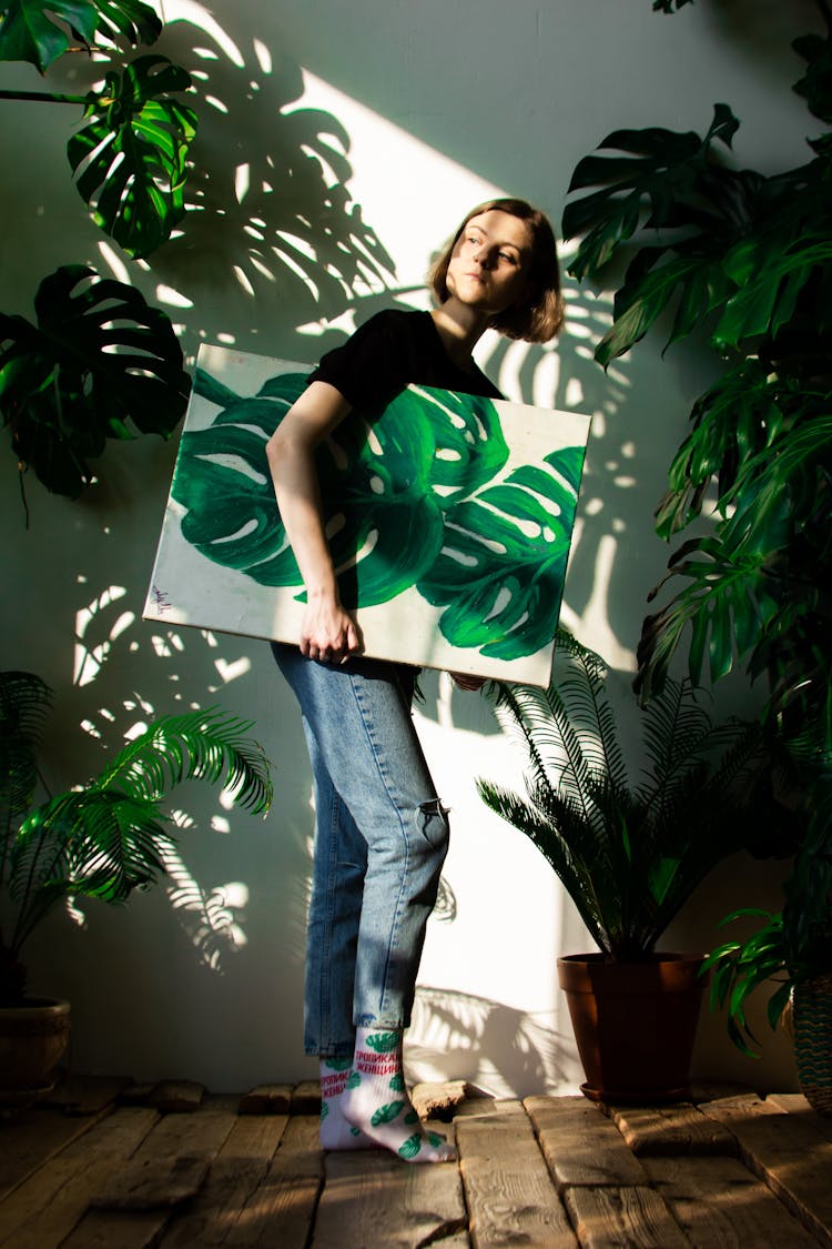 Woman Standing Between Monstera Plants And Holding A Painting Of Monstera Plant