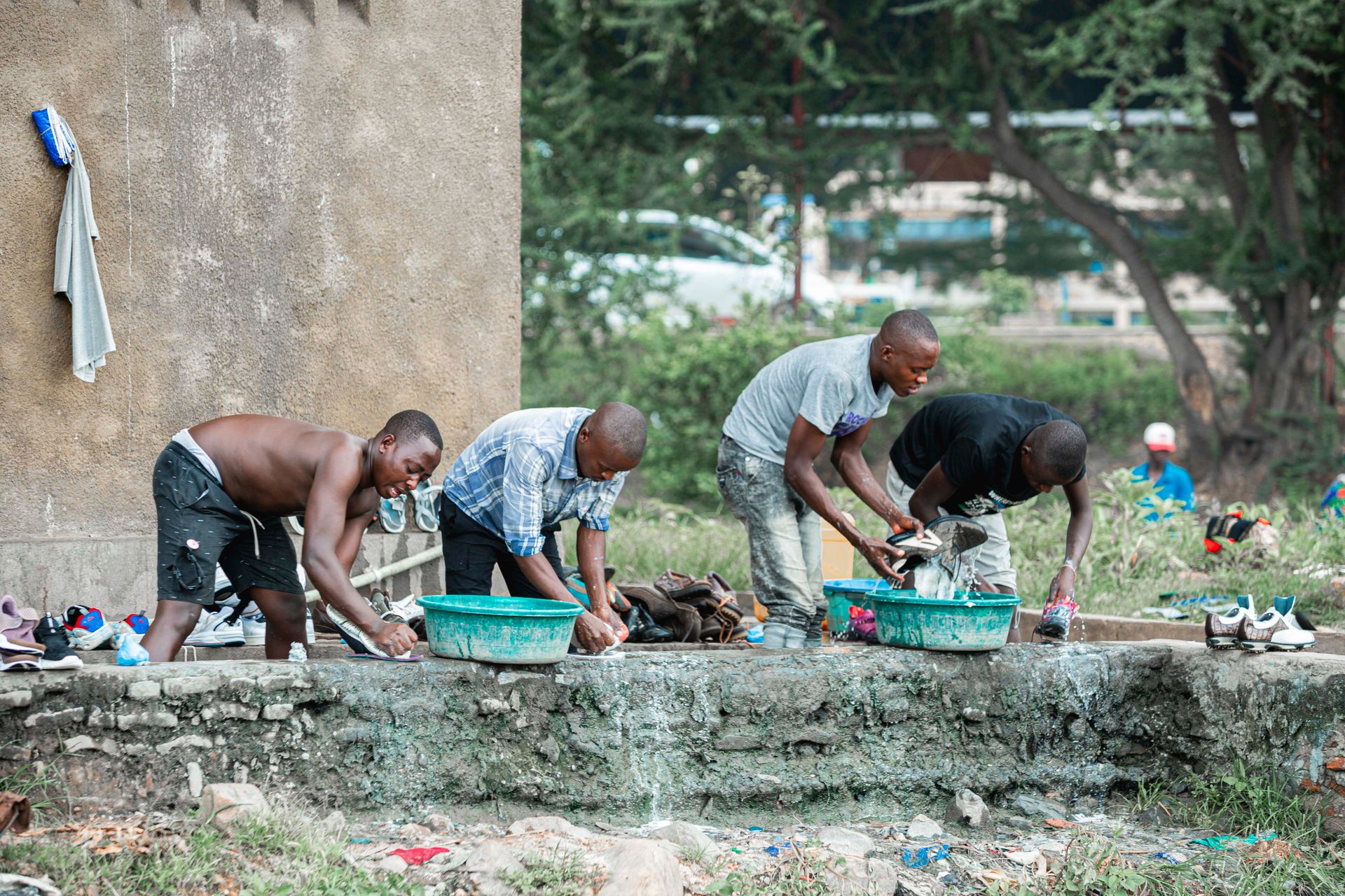 Four men washing clothes outdoors, using basins for cleaning shoes and clothing near a concrete wall.