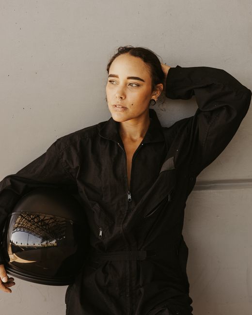 Portrait of a female motorsport racer in a black racing suit and helmet, leaning against a wall.