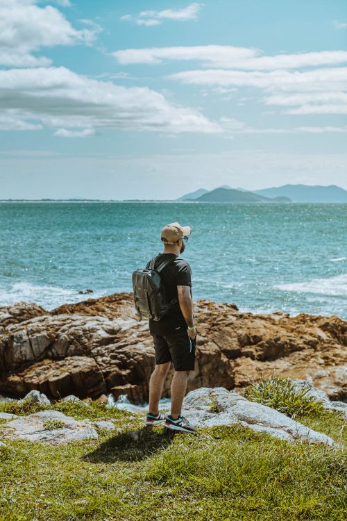 Free A man stands on a rocky cliff, gazing at the ocean in Garopaba, Brazil under a bright sky. Stock Photo