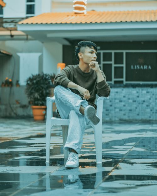 Free Asian man sitting thoughtfully on a chair in an outdoor setting on a rainy day. Stock Photo