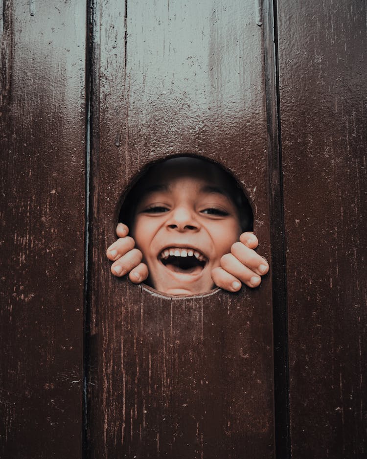 Laughing Child Face Looking Through Hole In Wooden Fence