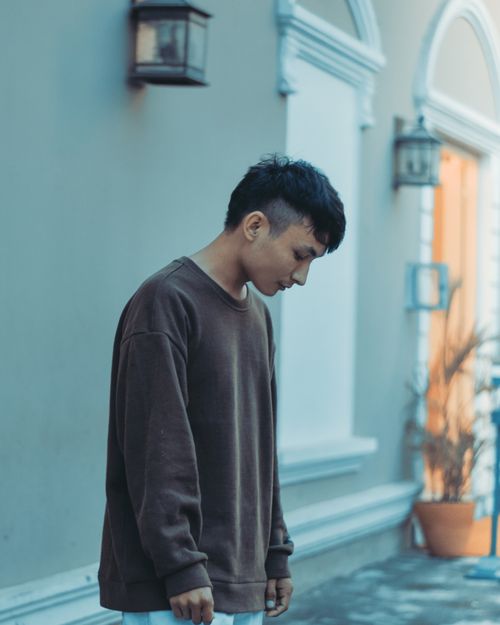 Free Young man wearing a brown sweater, looking down pensively against a blurred architectural background. Stock Photo