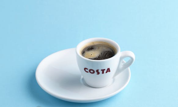 Close-up of a Costa coffee cup and saucer with dark coffee against a light blue backdrop.