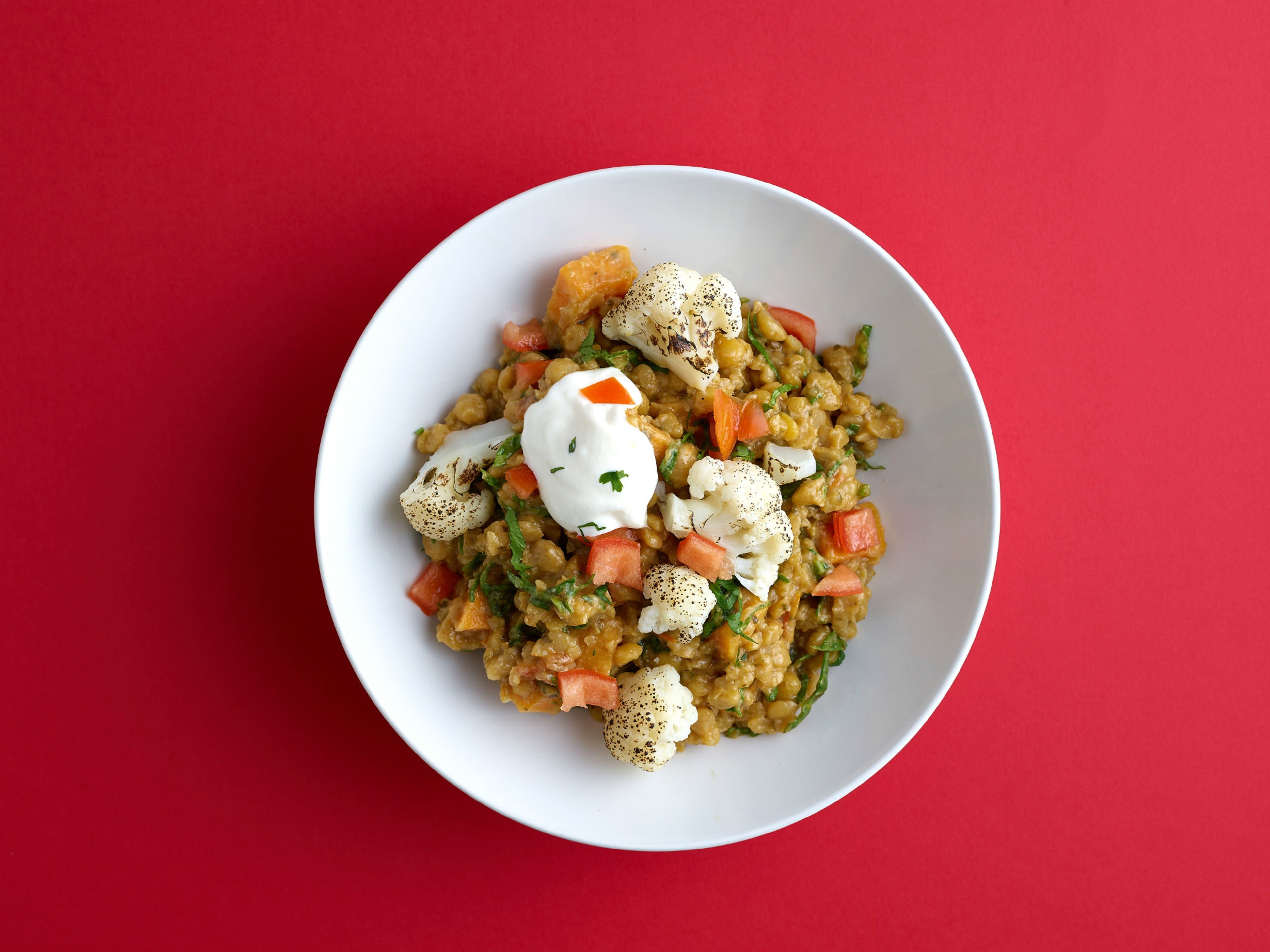 A white ceramic bowl filled with thick, russet-colored lentil stew, topped with a generous dollop of creamy white yogurt, fresh green mint and parsley, and a golden drizzle of olive oil