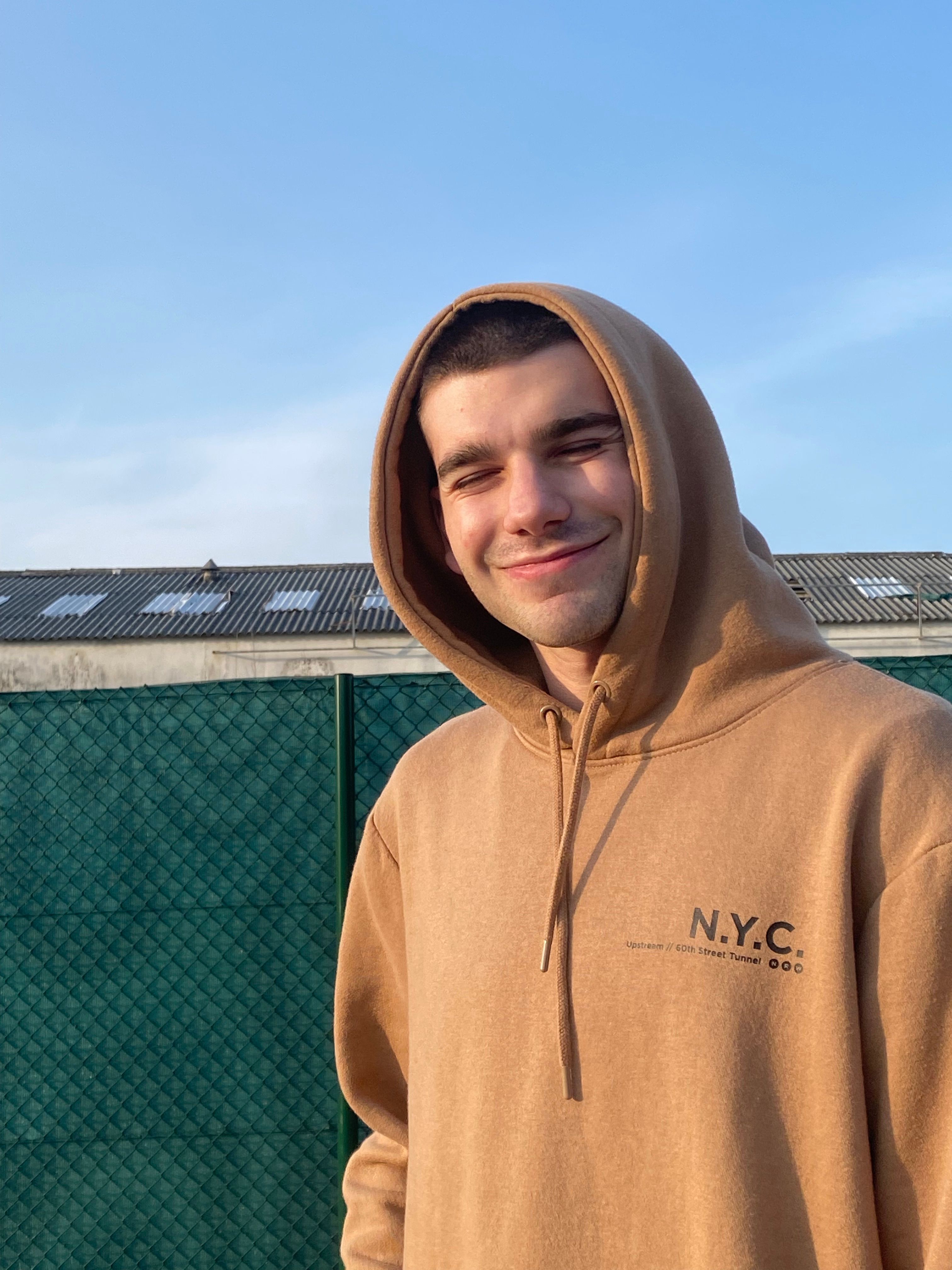 A young man smiling in an outdoor setting, wearing a tan hoodie with NYC text, under a clear blue sky.