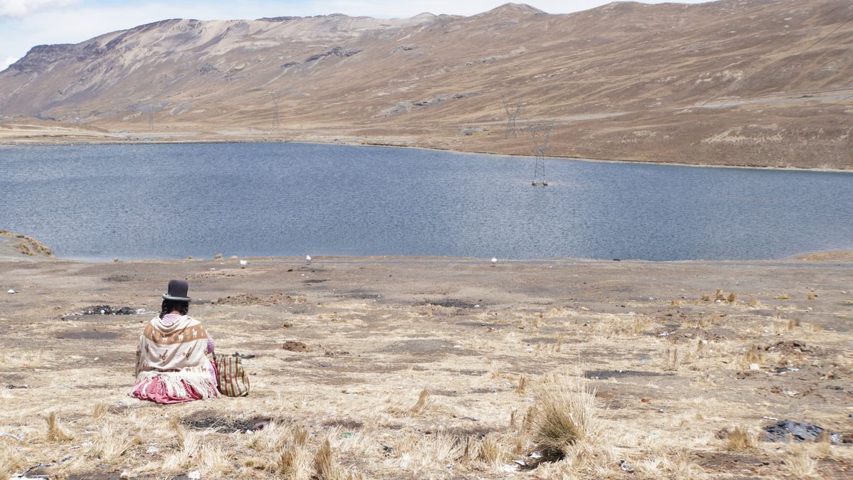 A woman in traditional Andean dress sitting on the shore of a still lake in the Bolivian highlands with bare hills behind