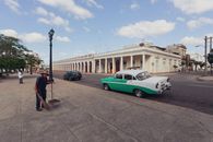A street cleaner in a hat sweeping the pavement near a vintage car and colonial building.
