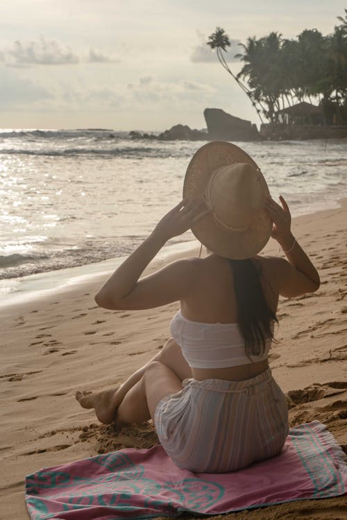 Free A Woman in White Tube Top Sitting on the Beach while Wearing a Hat Stock Photo