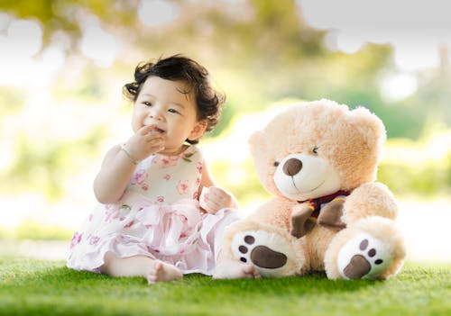 Baby Sitting on Green Grass Beside Bear Plush Toy at Daytime