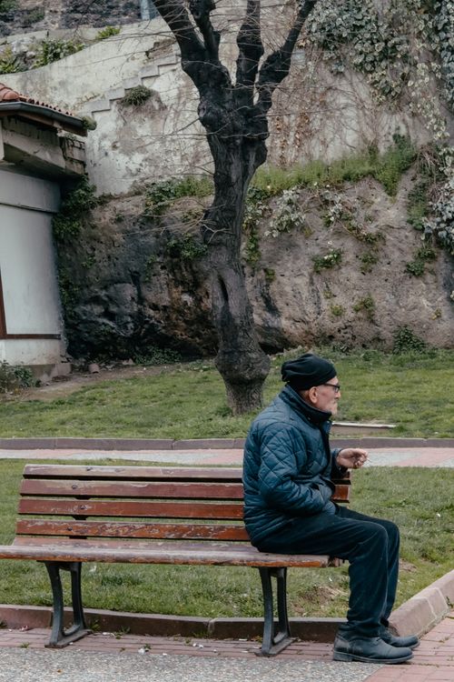 Free An elderly man in a winter jacket sits on a park bench, enjoying a peaceful moment outdoors. Stock Photo