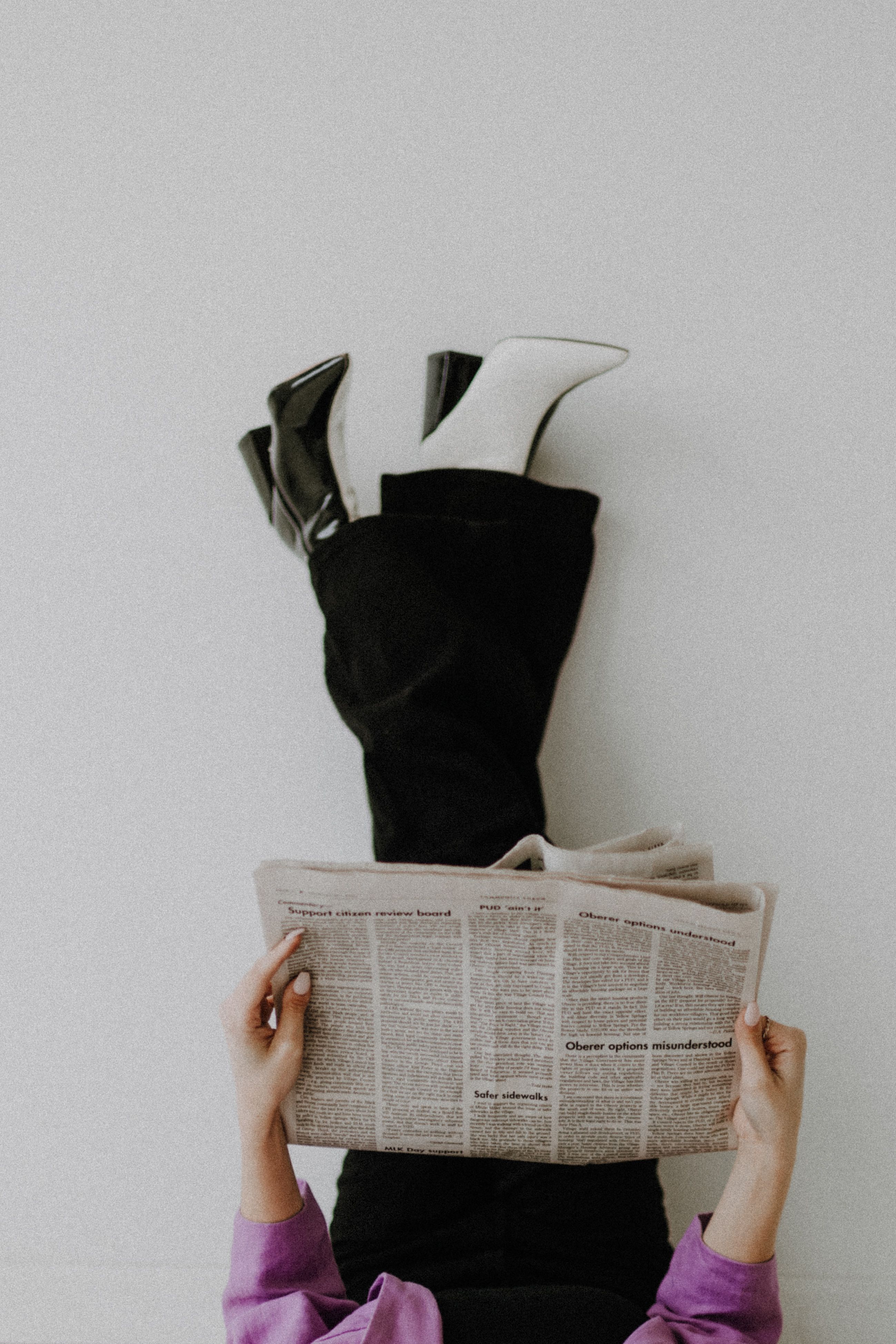 Fashionable woman reading newspaper with legs up against a wall in a studio setting.