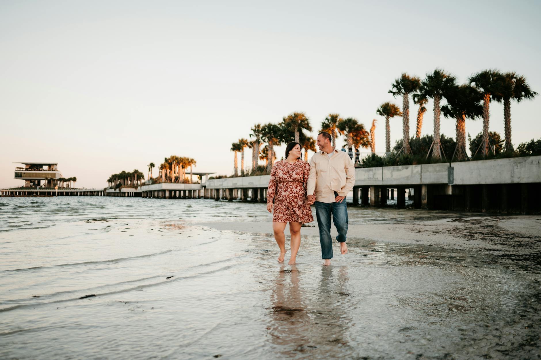 A couple enjoys a romantic walk on a St. Petersburg beach with palm trees lining the shore at sunset.