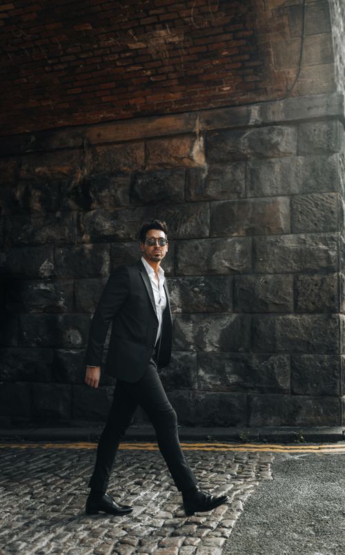 Free Confident man in black suit walking under a brick archway in an urban setting. Stock Photo