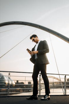 Stylish man in suit checks phone on urban bridge during sunset.
