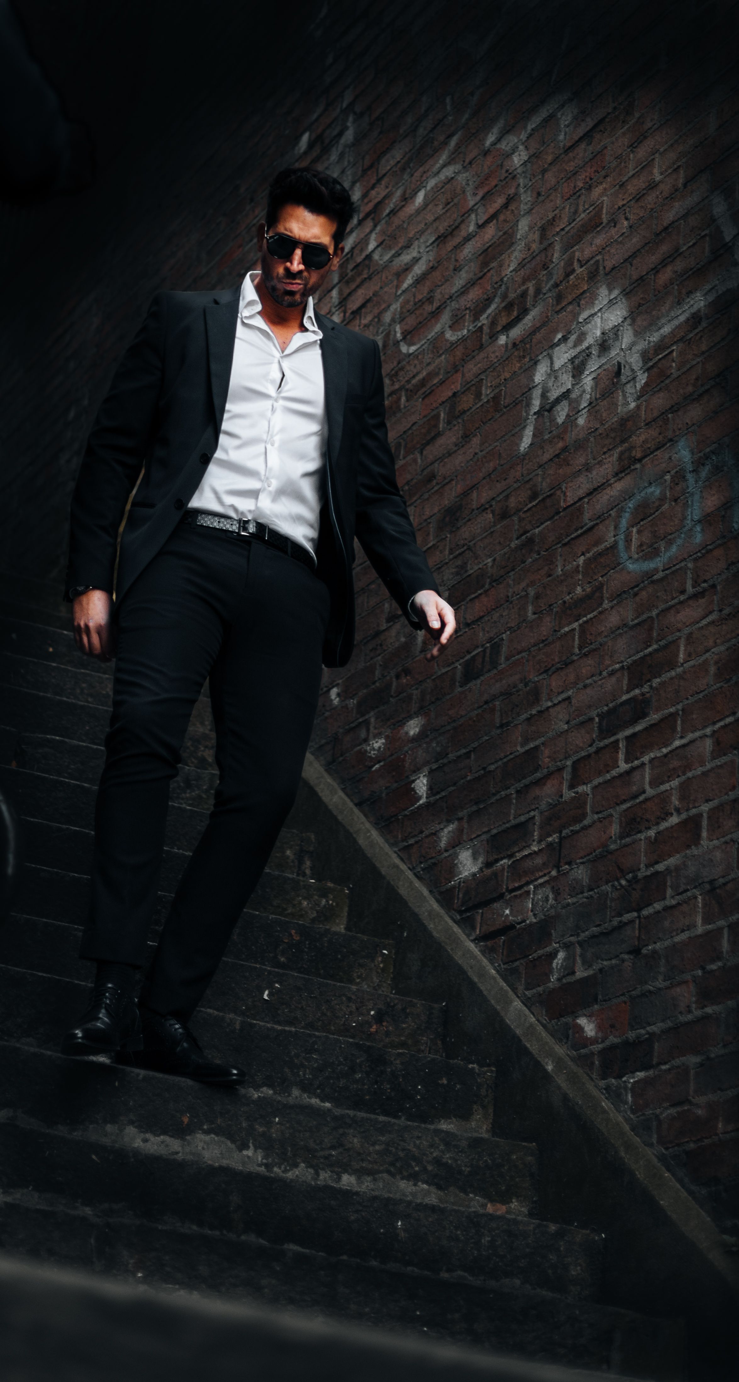 A fashionable man in sunglasses walks down an urban brick staircase, exuding elegance.