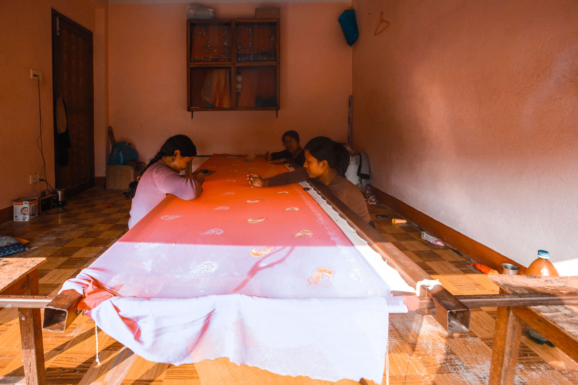 Women involved in intricate embroidery work on long fabric pieces in a workshop setting.