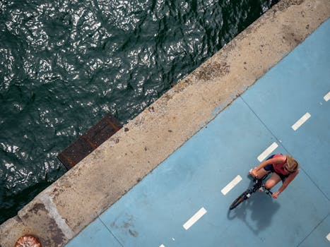 Aerial image of a woman cycling on a blue riverside path, showcasing a vibrant outdoor scene.