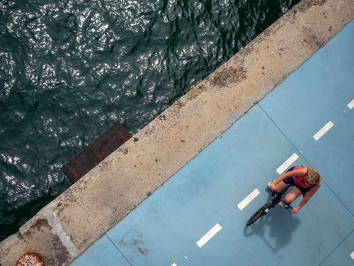 Free Aerial image of a woman cycling on a blue riverside path, showcasing a vibrant outdoor scene. Stock Photo