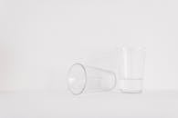 白色表面上放着一对透明玻璃杯，一个直立着装有水，另一个侧放在旁边