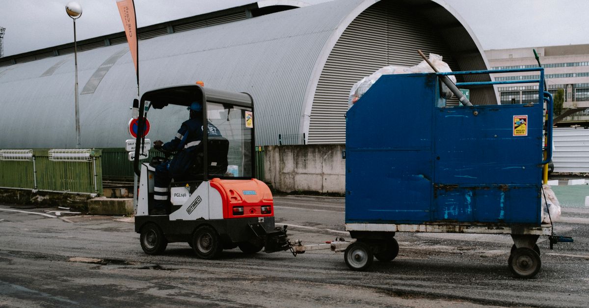 Forklift operator pulling a blue container outdoors near industrial facility.