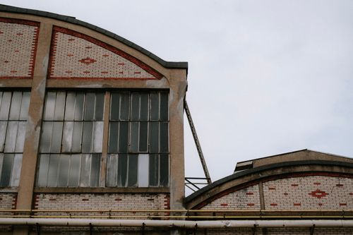 Free Close-up of a red brick factory building with large, weathered windows under a cloudy sky. Stock Photo
