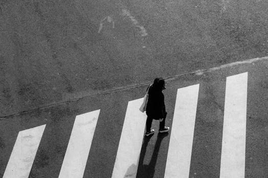 A woman crossing an urban zebra crossing in a black and white cityscape captured from above.