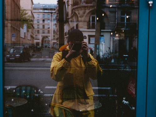 Free A photographer captures a city reflection through a window, wearing a yellow jacket. Stock Photo