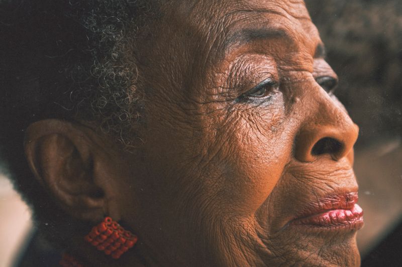 Close-up profile portrait of an older Black woman with gray curls and red beaded earrings, her open eye lit by warm daylight as she looks into the distance.