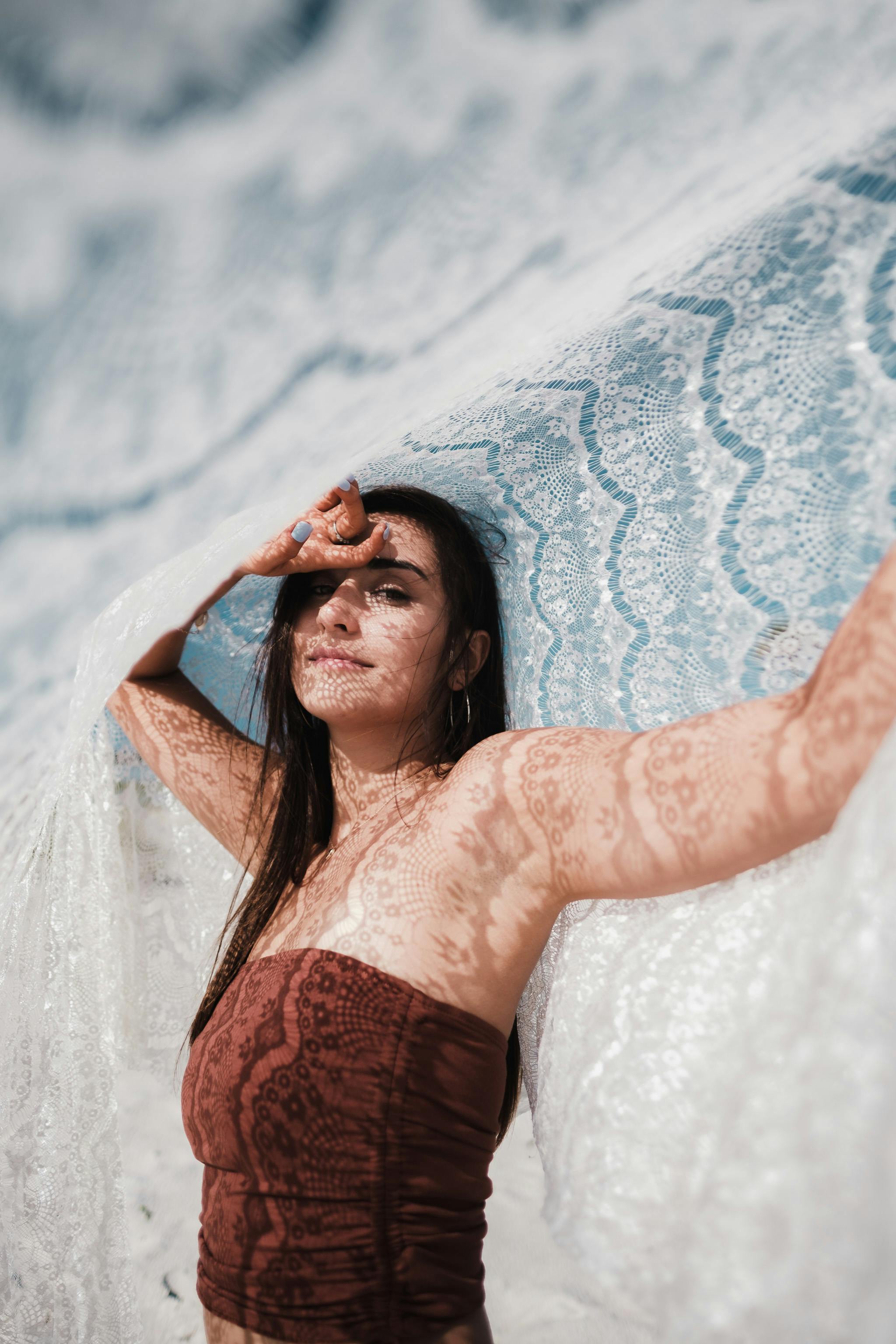 A woman poses gracefully outdoors, draped in lace, creating intricate light patterns on her skin.